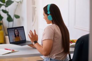 A woman wearing headphones is at a desk, using a laptop and talking to another woman, suggesting she is engaged in an online task or conversation."