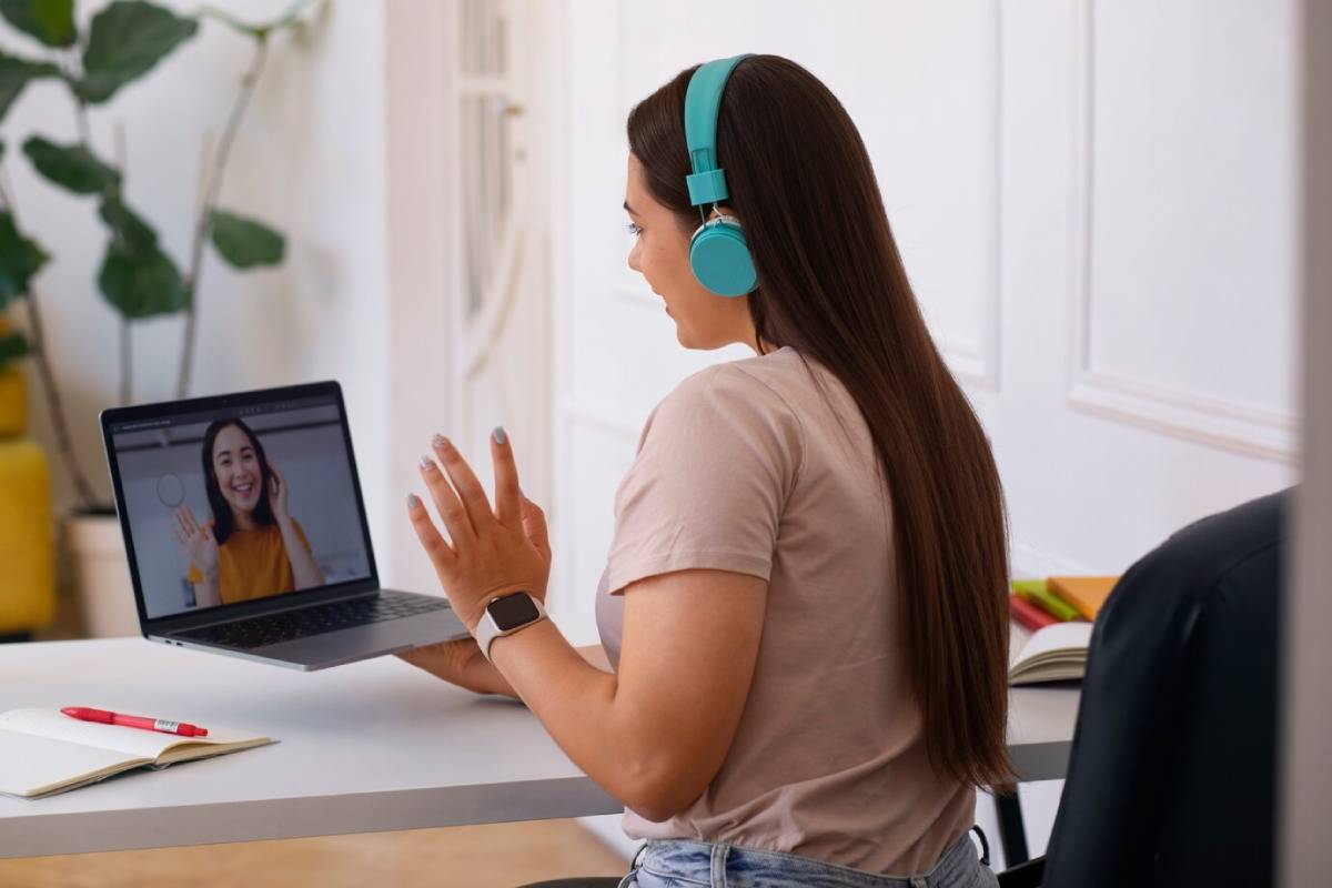 A woman wearing headphones is at a desk, using a laptop and talking to another woman, suggesting she is engaged in an online task or conversation."