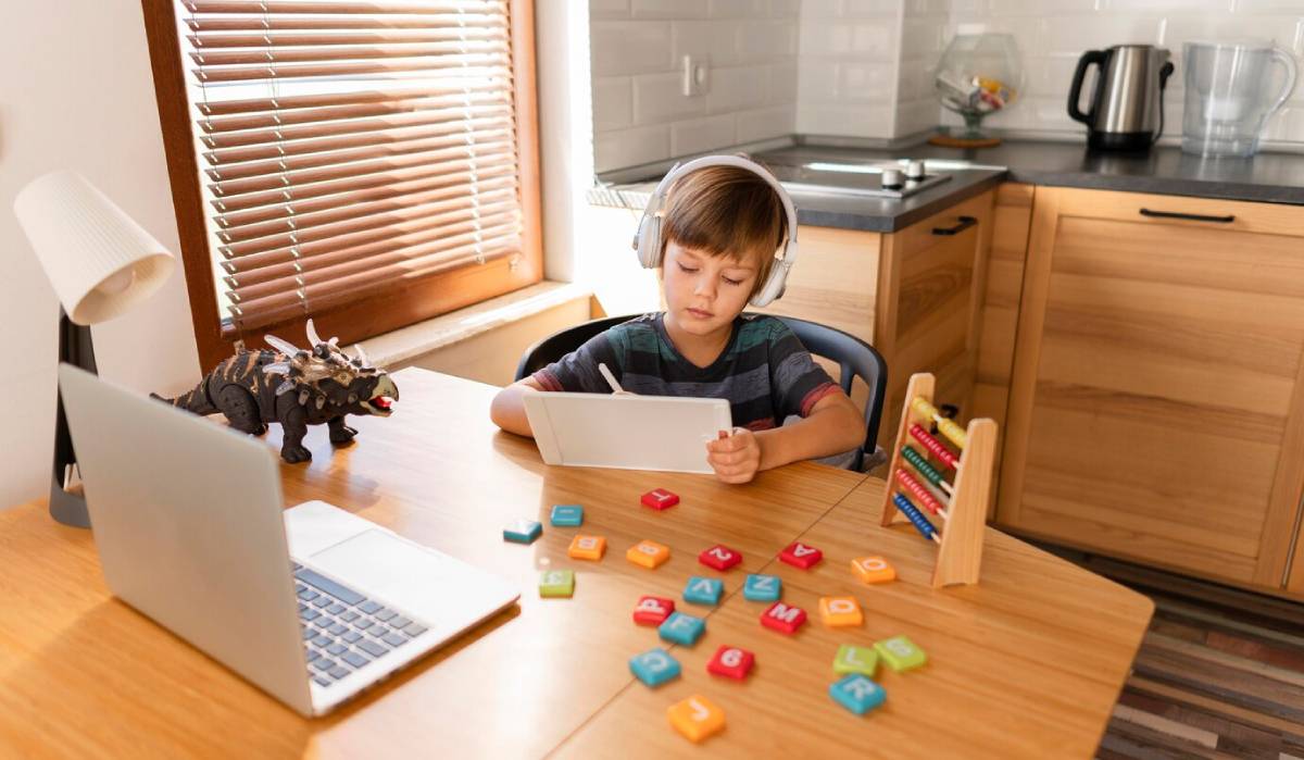 A young boy at a wooden table, engaged with his laptop, surrounded by game based learning stuff. 