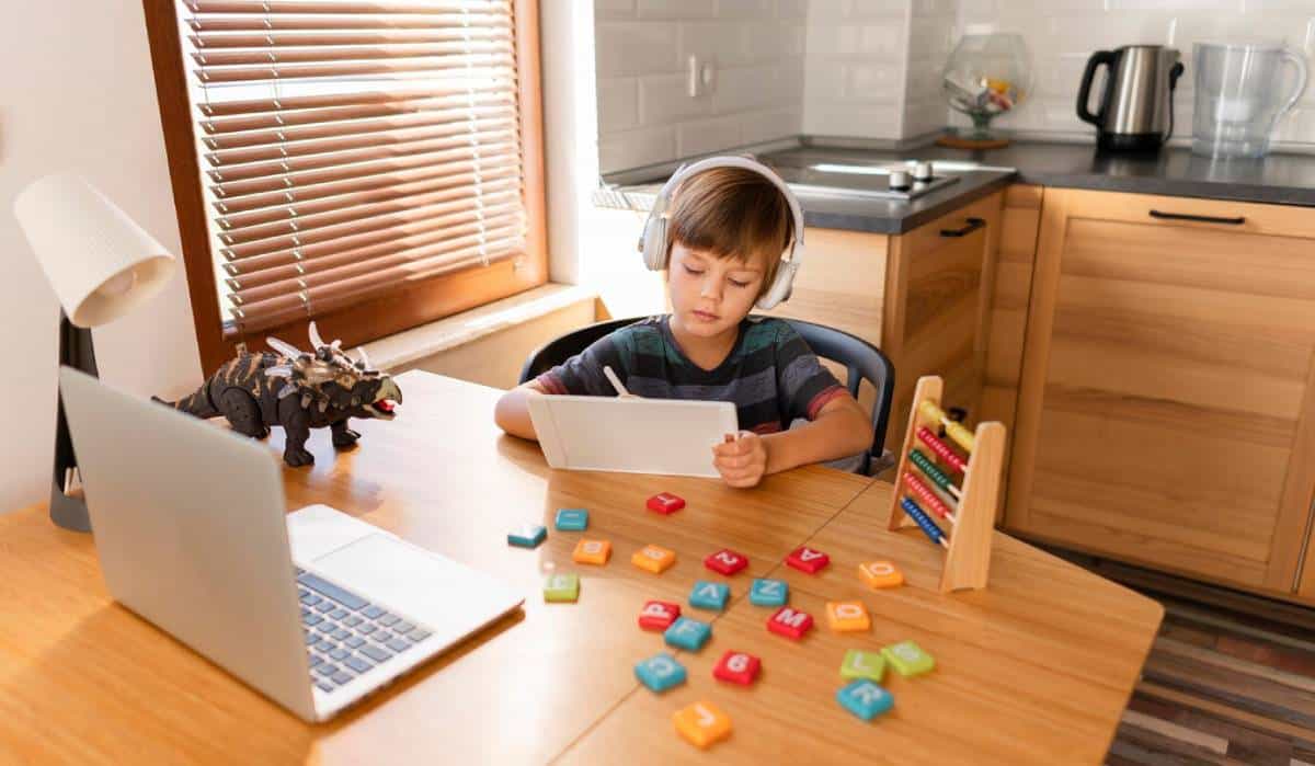 A young boy at a wooden table, engaged with his laptop, surrounded by game based learning stuff.
