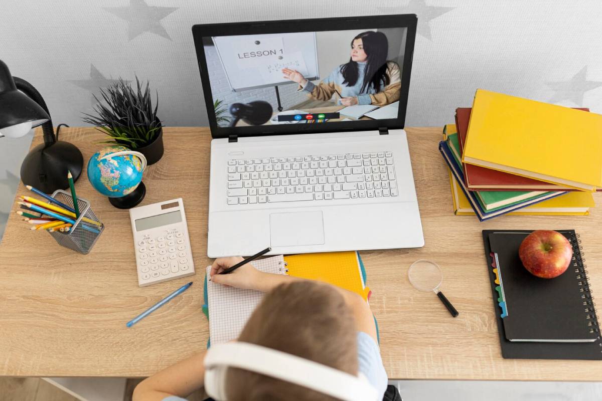  A child sits at a desk, focused on a laptop, with an open book beside them.