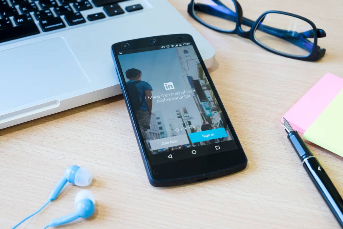 A smartphone displaying the LinkedIn login screen, positioned near a laptop, glasses, headphones, and sticky notes on a wooden desk. 