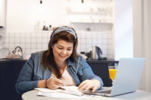 A woman sits at a table with a laptop open and a pen in her hand, focused on her work.