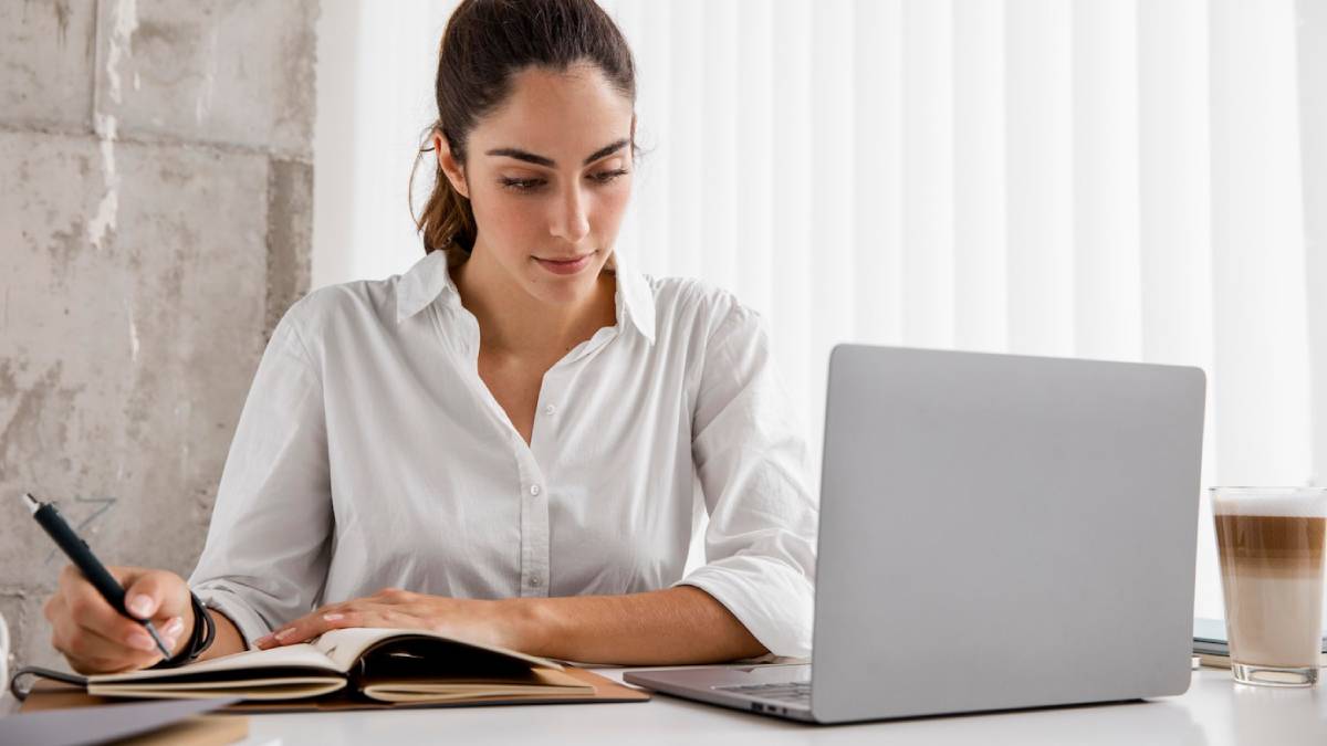 A woman sits at a desk, focused on her laptop, with a pen in hand, ready to take notes or write.