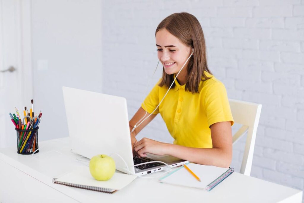 A girl in a yellow shirt sits at a desk, focused on her laptop while working or studying.