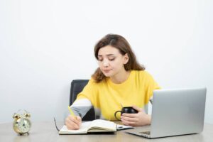A woman sits at a desk with a laptop open and a cup of coffee beside her, focused on her work.