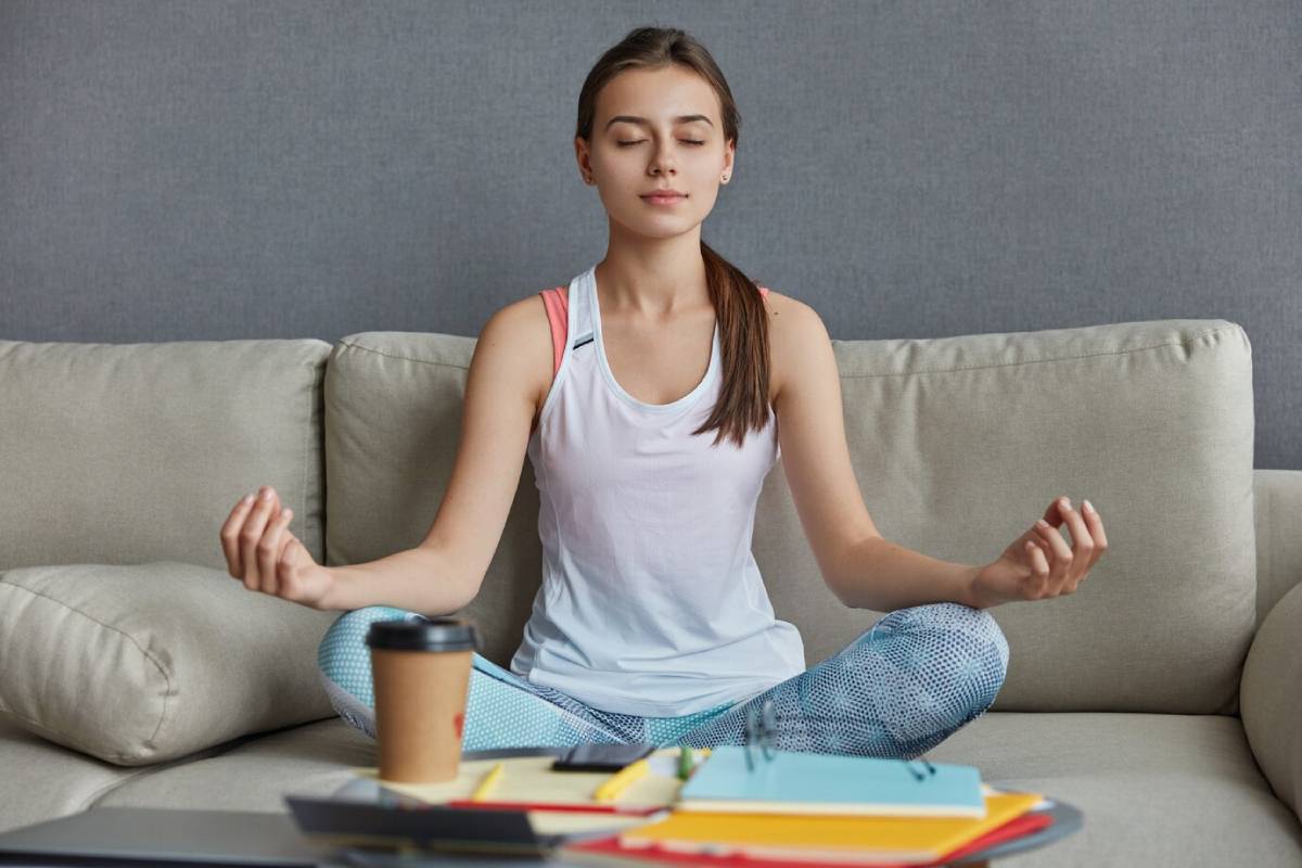 A woman sits cross-legged on a couch, meditating peacefully in a yoga position.
 
