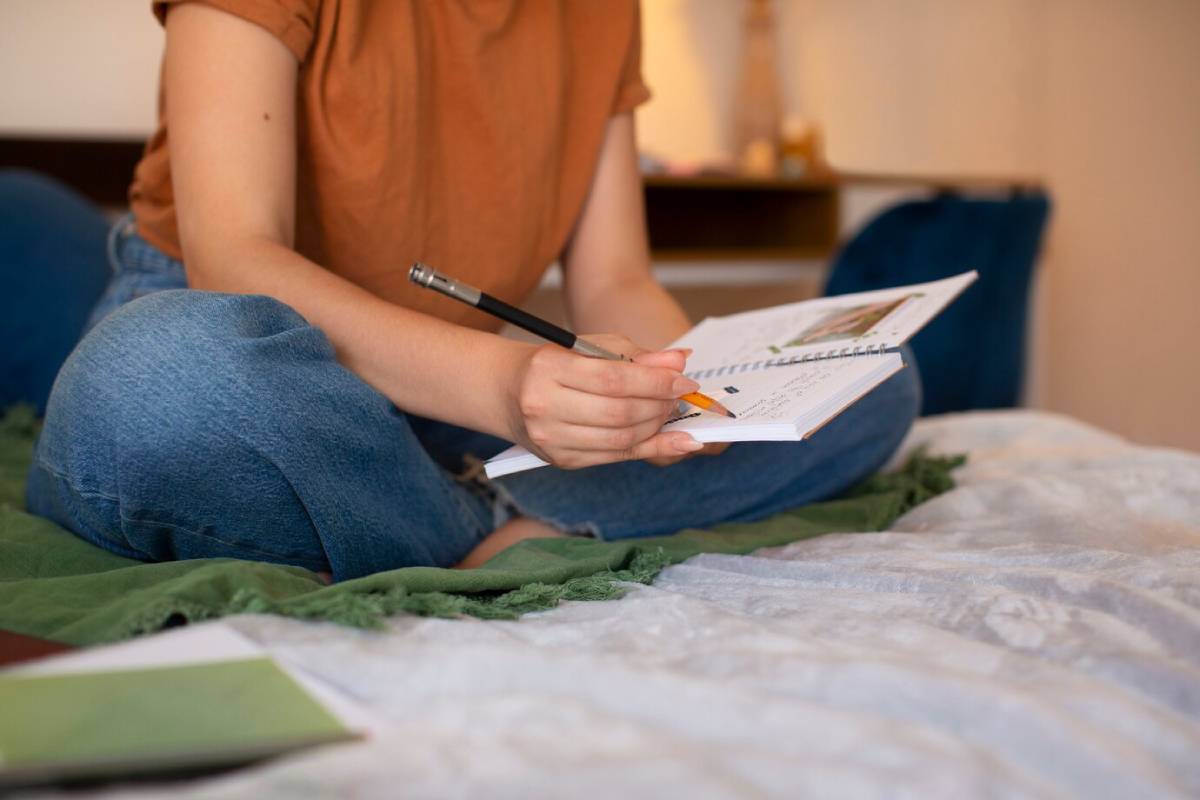 A woman sits on a bed, writing in a notebook with a pen, surrounded by a cozy bedroom setting. 