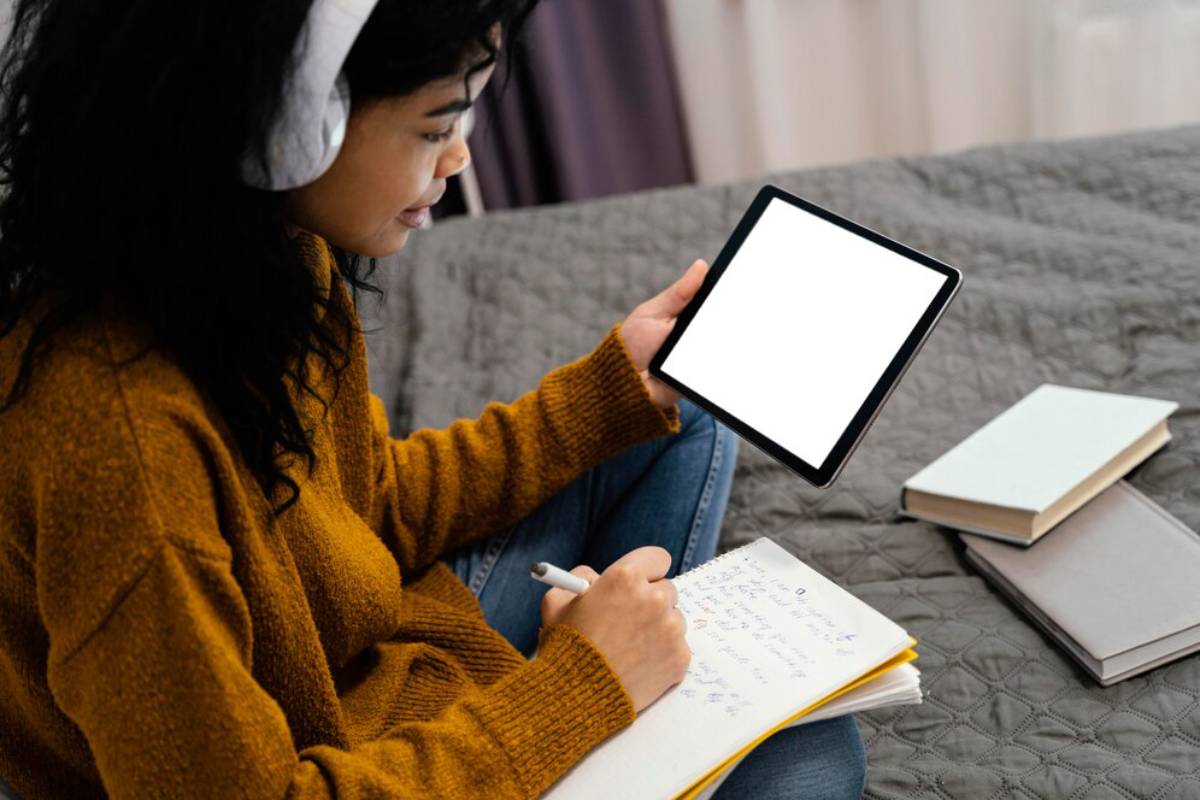 A woman sits on a bed wearing headphones, focused on a tablet in her hands and writing notes. 