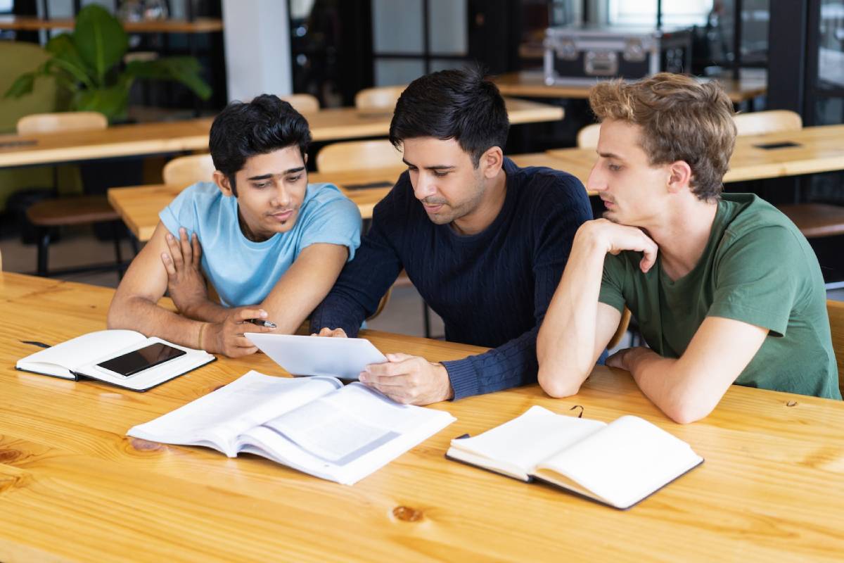 Three young men seated at a table, focused on a tablet in front of them.
 