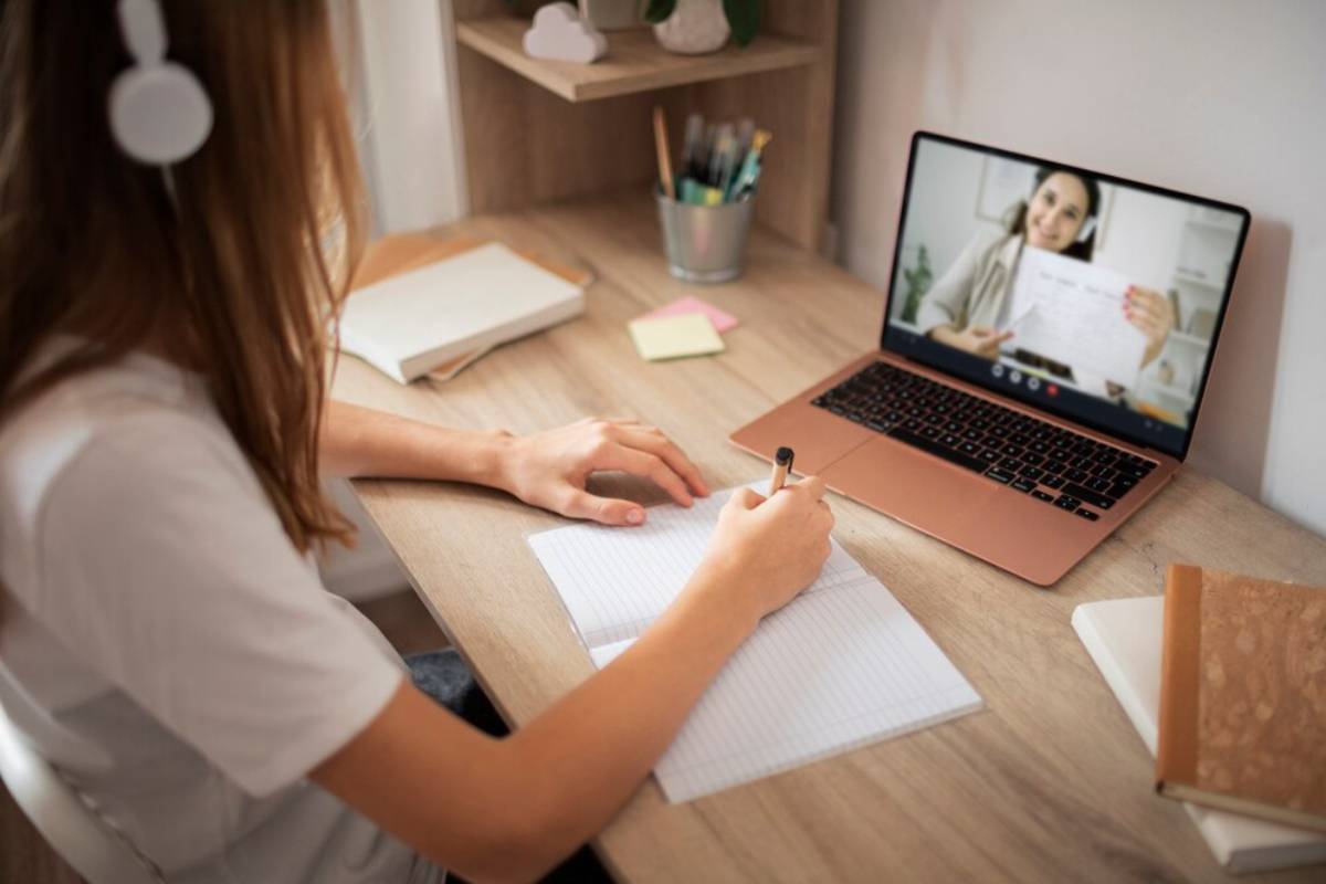 A woman is watching a video lecture and writing notes in a notebook.