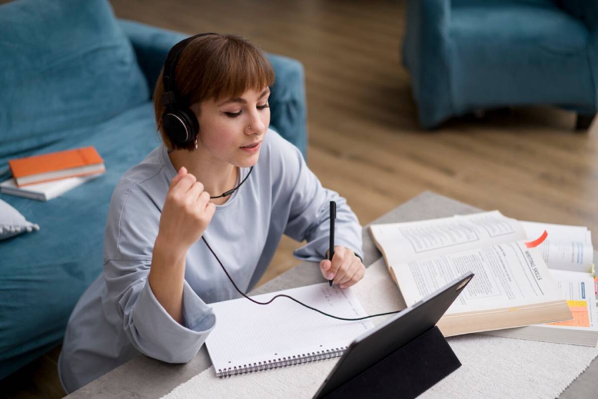 A woman with headphones sits at a table, focused on her laptop and taking notes in a notebook.