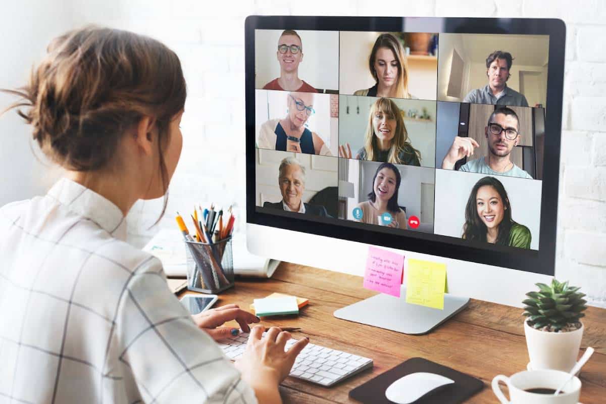 A woman sits at a desk, focused on a computer screen displaying a video call with multiple participants.