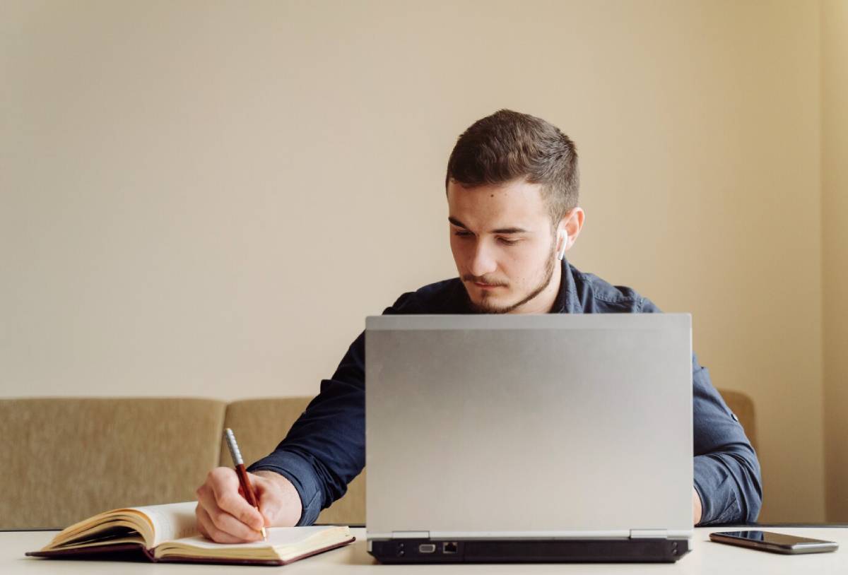 A man seated at a table, working on a laptop with a pen in hand, focused on his task.
