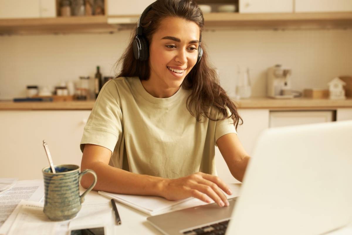 A woman with headphones sits at a table, focused on her laptop, engaged in work or a virtual meeting. 