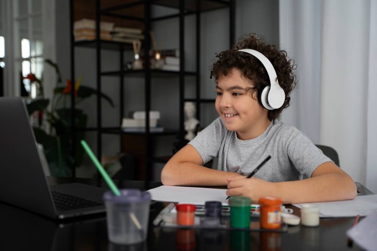 A boy with headphones sits at a table, focused on his laptop while engaged in an activity. 