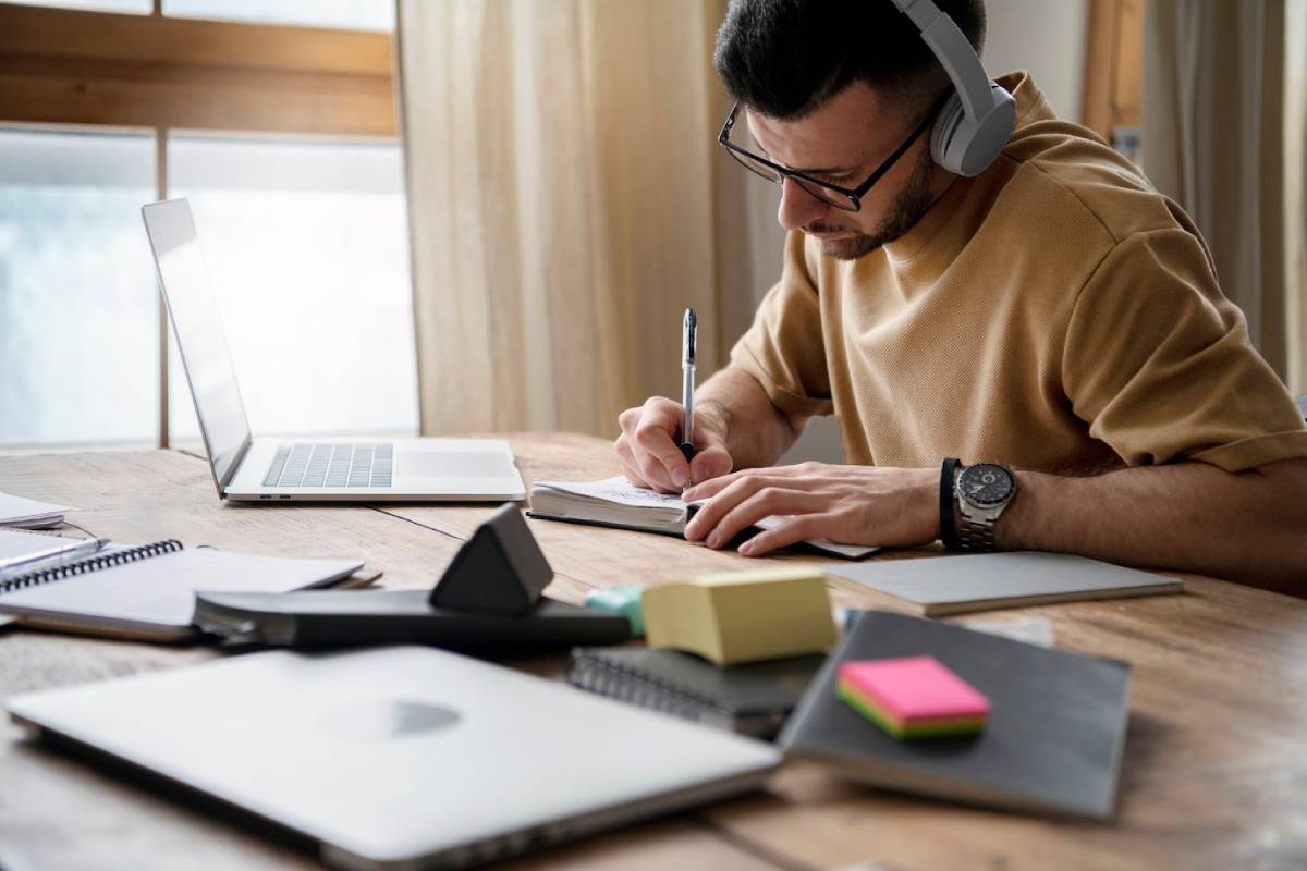 A man wearing headphones sits at a desk, focused on writing in a notebook.