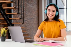 A woman in a bright orange shirt wears teal headphones while studying at a modern desk with a laptop and notebooks.