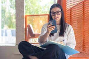 A person sits cross-legged, holding a phone and notebook, in a bright, modern space with orange accents and green foliage outside.