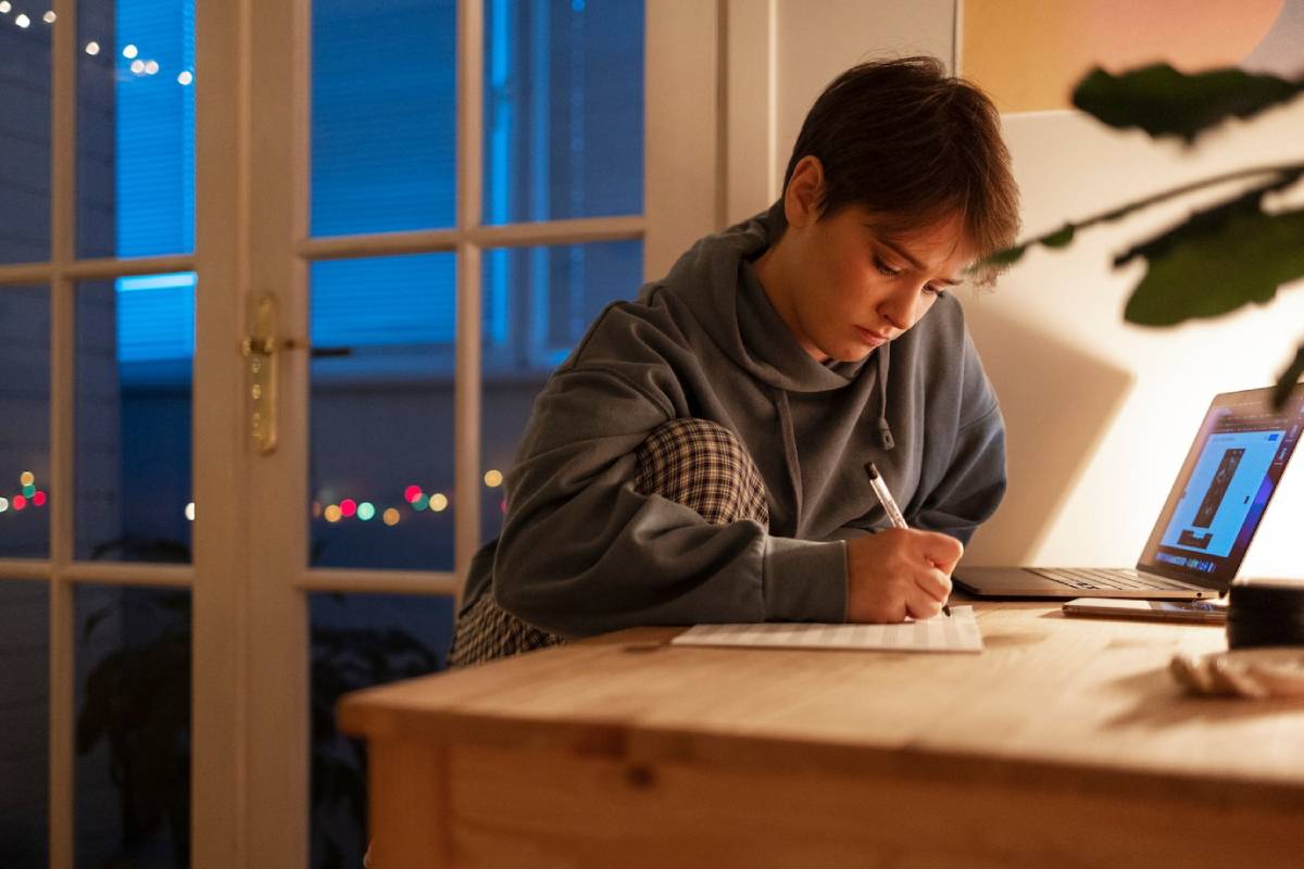 A person writes on a notepad at a wooden table, with a laptop and soft, decorative lights glowing in the background. 