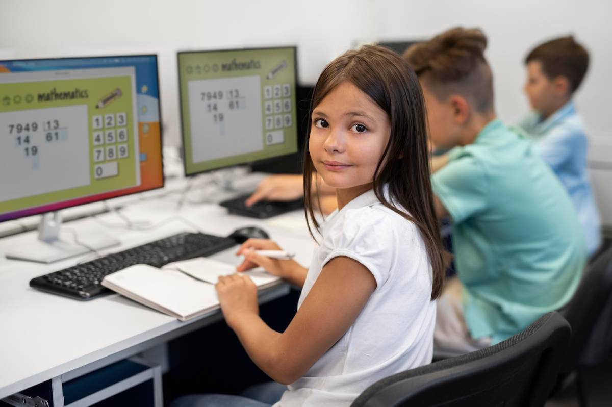 Students engaged in a mathematics lesson on computers, with notebooks open and math problems displayed on their screens. 