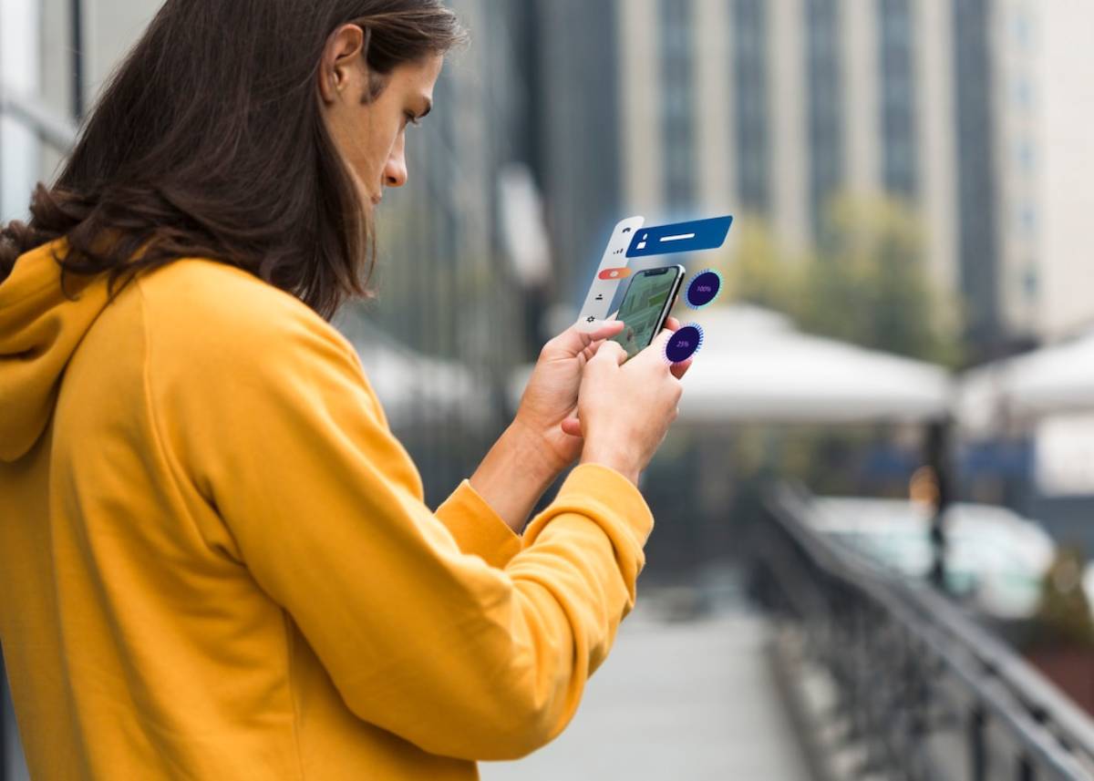 A person in a yellow hoodie using smartphone, with modern buildings in the background and outdoor seating. 