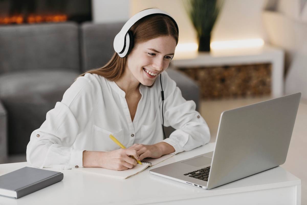 A person wearing headphones sits at a desk, writing in a notebook beside a laptop, in a cozy, well-lit room. 