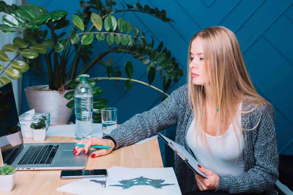 A woman with long hair sits at a desk, using a laptop and holding a clipboard, surrounded by plants and a water bottle.