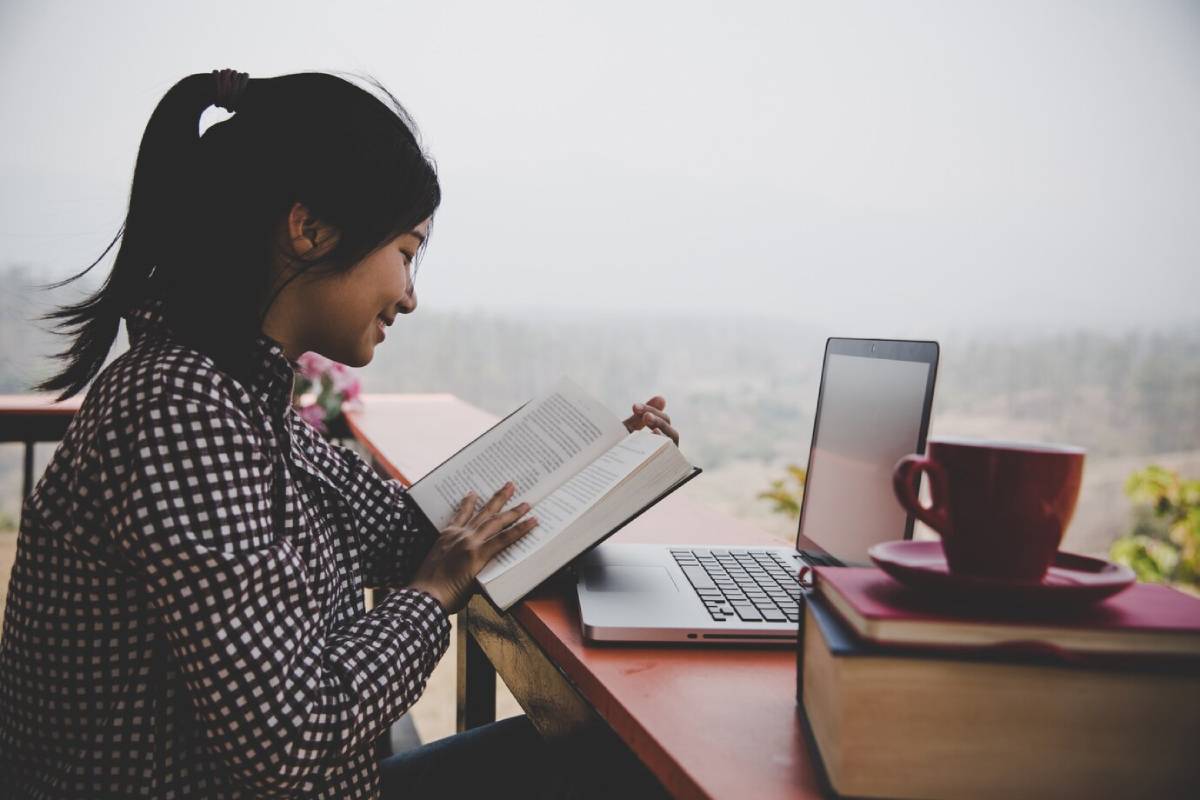 A person reads a book at an outdoor table, with a laptop and a coffee mug beside them, overlooking a scenic landscape. 