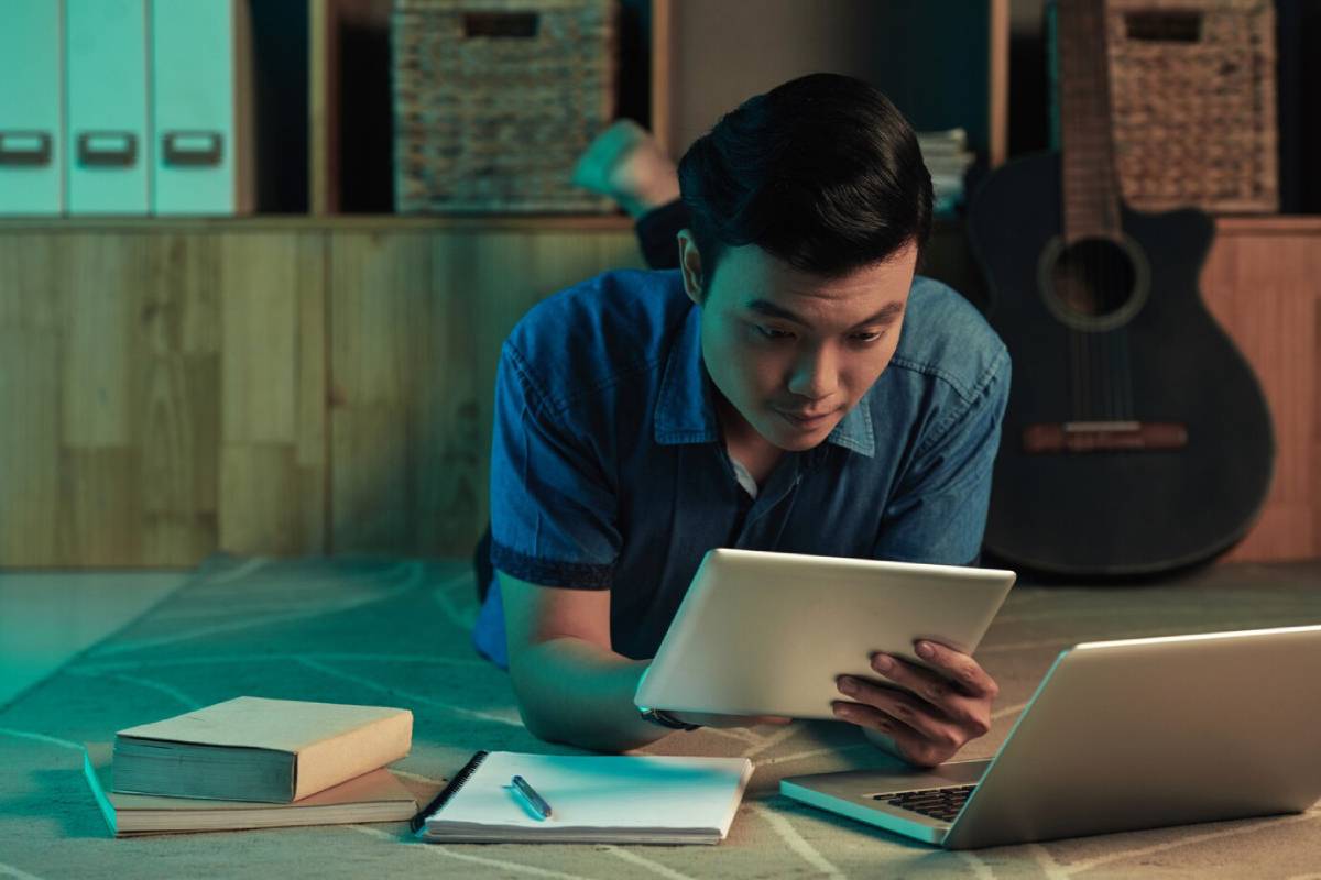 A person lies on a rug, using a tablet, with an open laptop and notebooks nearby, surrounded by books and a guitar in the background. 