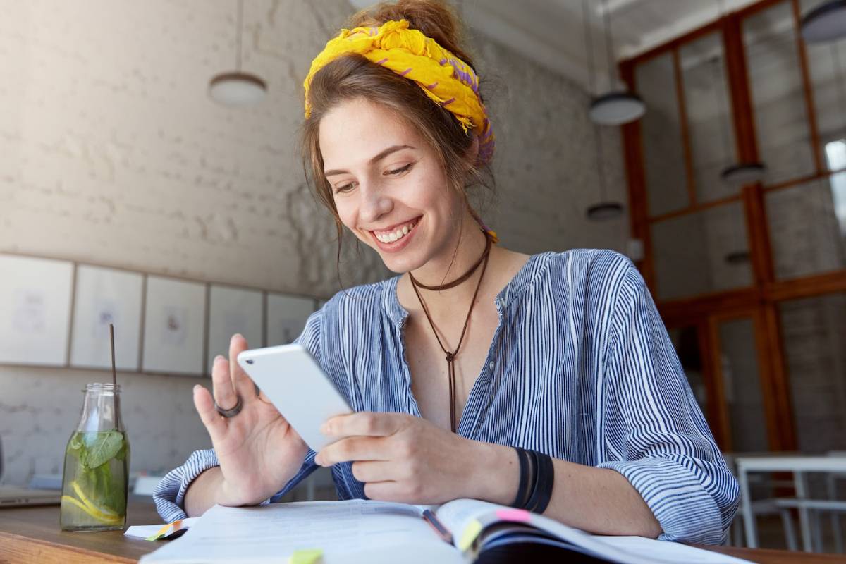 A person in a striped shirt uses a smartphone while studying at a table, with a glass of mint drink and open notebooks nearby.